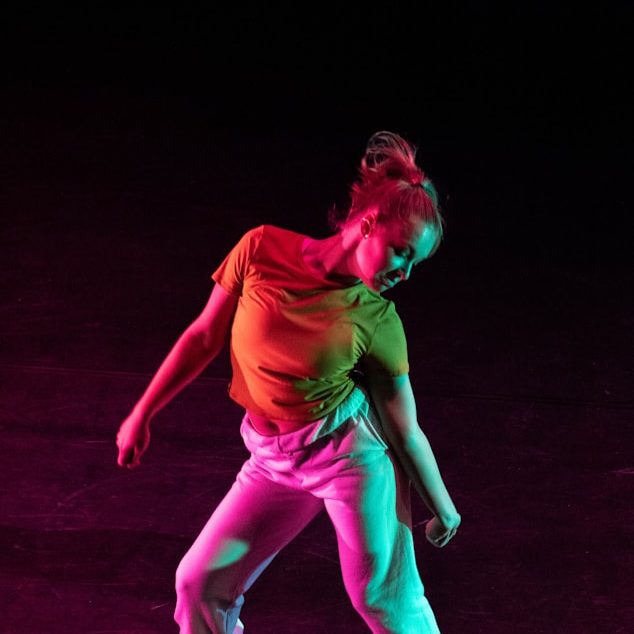 Dancing Artist Tänzerin in bunter Kleidung, die dynamisch auf einer dunklen Bühne bewegt.